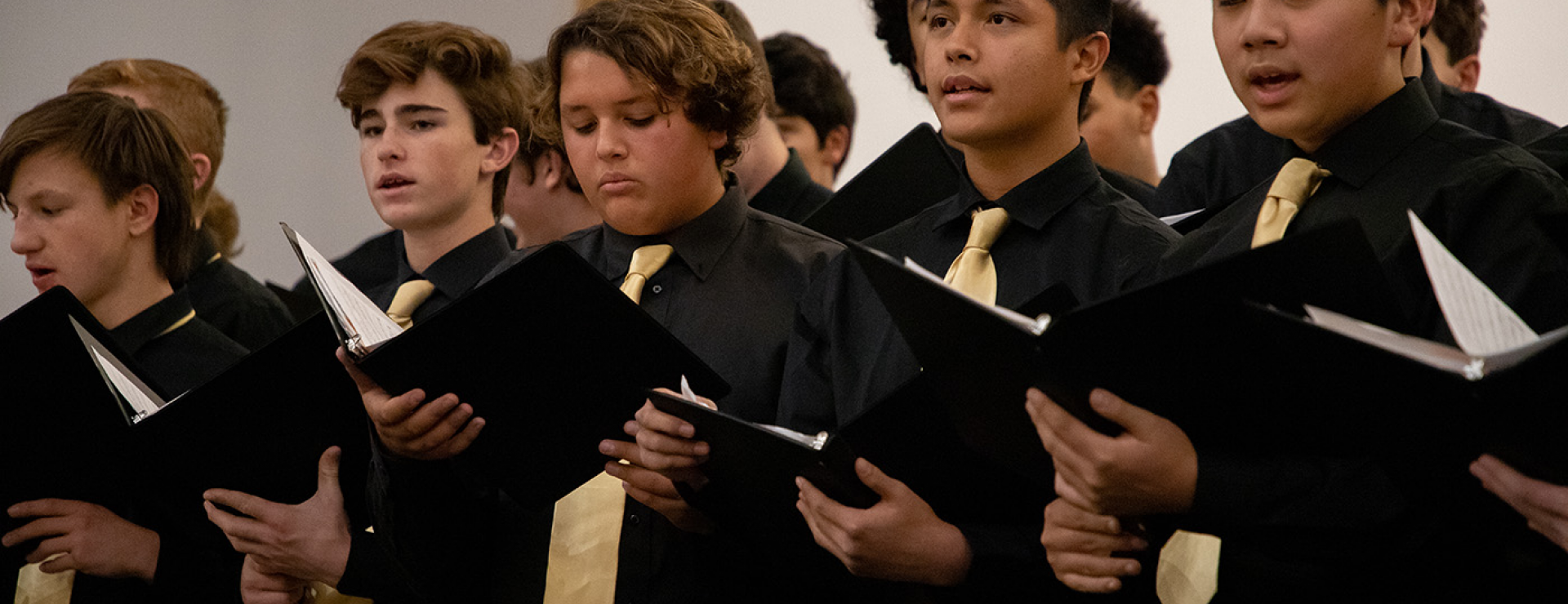 Row of boys singing at a concert. Wearing all black with gold ties and holding song books. 