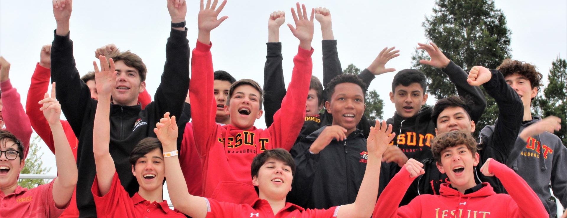 small crowd in stands all in Jesuit logo apparel arms raised high cheering 