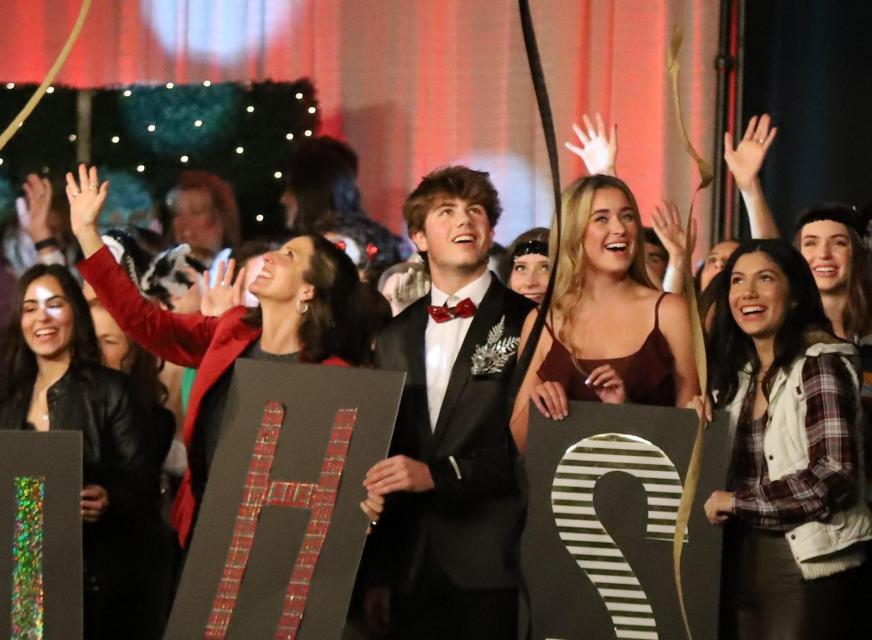 Stage of festive students in formal gear holding JHS signs while streamers fall.