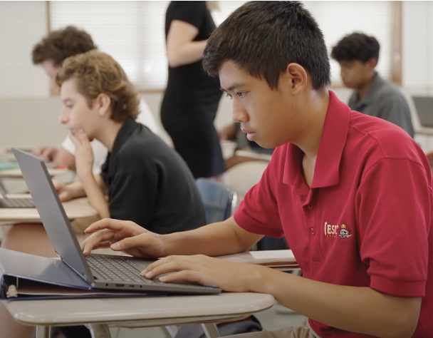 close up of a student typing on his computer, at a desk in a classroom setting