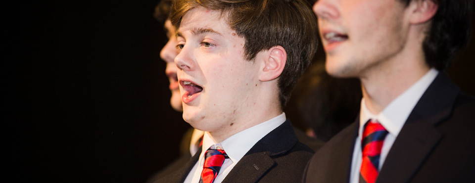Closeup of students in black suits and red ties with mouths open in song.