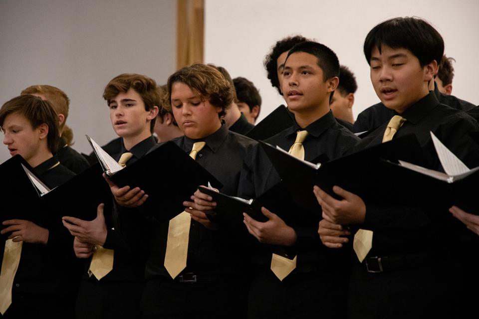 Row of boys singing at a concert. Wearing all black with gold ties and holding song books. 
