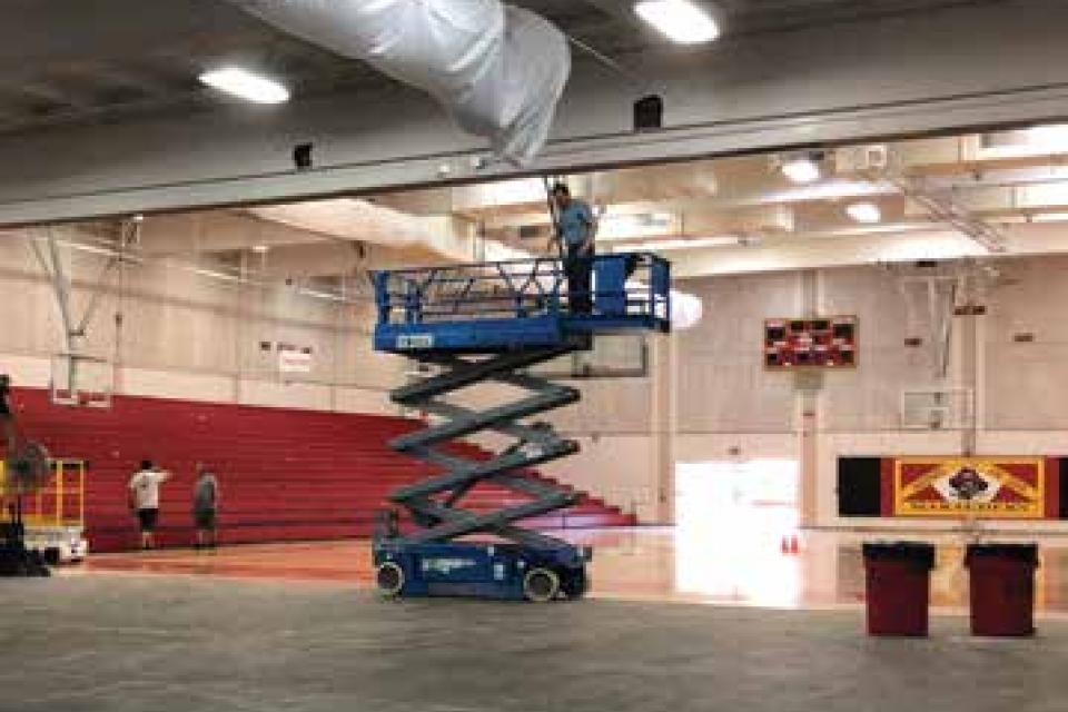 Scissor lift with man working on HVAC in the middle of the gym.