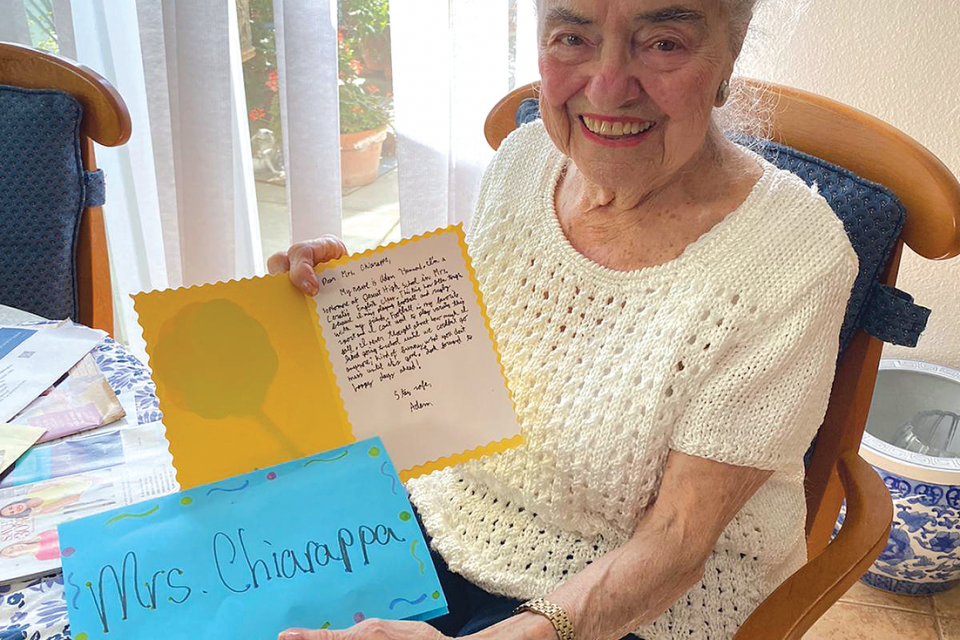 Elderly woman sitting and smiling while holding a card in each hand.