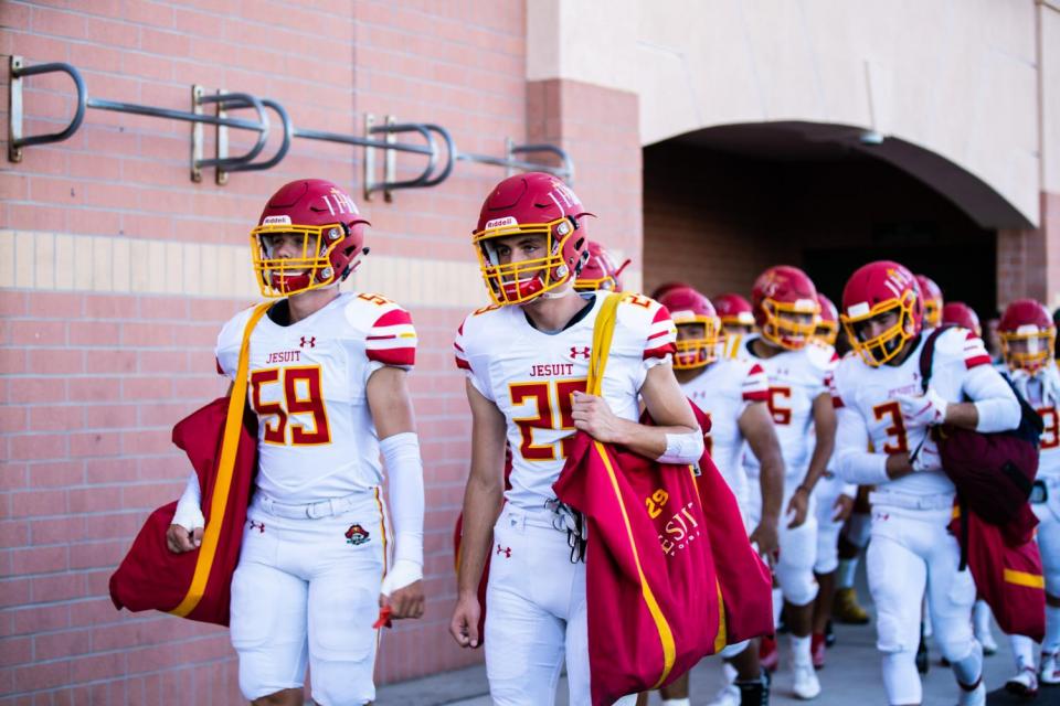 Line of football players in full uniform carrying bags of gear.