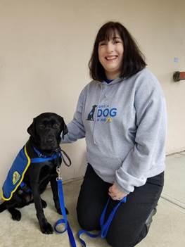Smiling Jennifer kneeling next to a sitting young dog wearing a service vest.