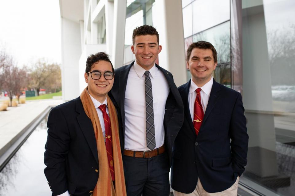 Three students smiling in formal dress  by the chapel.