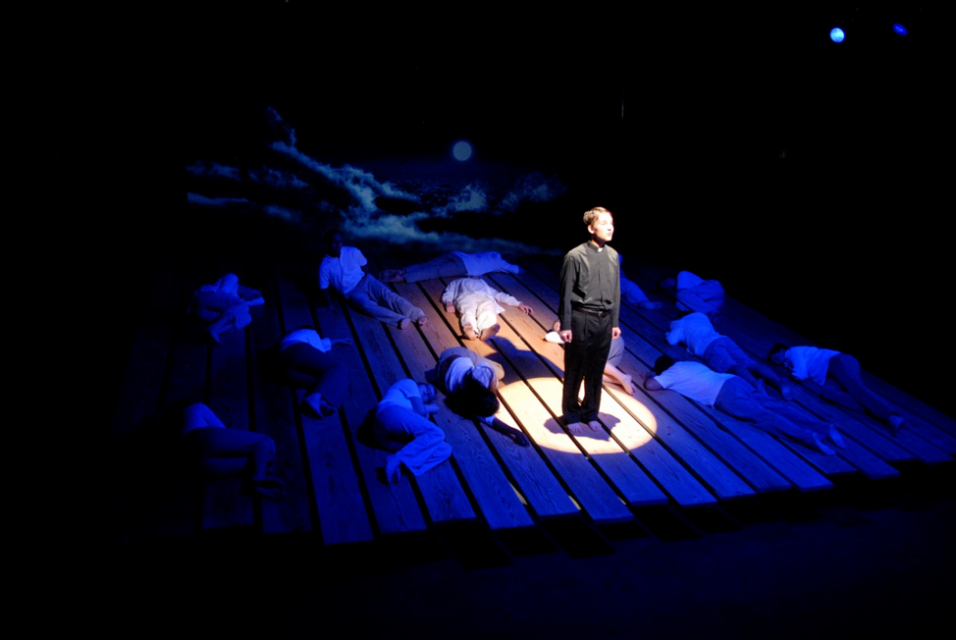 Dark stage with actors laying down on plank floors, while one student stands in spotlight.