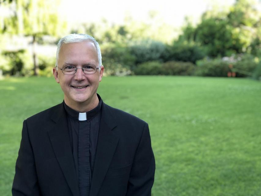 Image of Rev. Gilson, S.J. on campus in front of garden