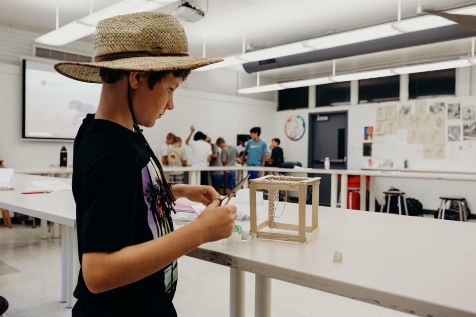 Image of young camper in a sun hat building a structure of sticks inside a classroom