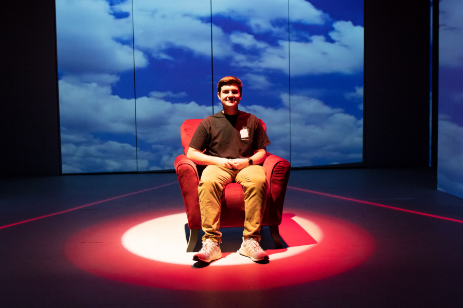 Student sitting alone on stage in red arm chair with dramatic lighting and staging.