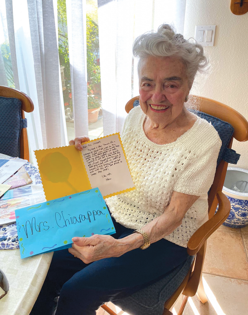 Elderly woman sitting and smiling while holding a card in each hand.